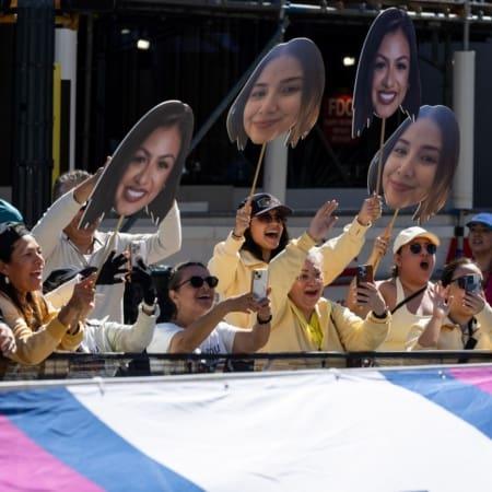 A group of excited people stand behind a barrier, cheering and holding large cutouts of a smiling woman's face. Many are taking photos with their phones, anticipating future trends for 2027, and most wear light-colored clothing and sunglasses. Austin Marathon Half Marathon & 5K