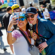 Two people smiling and taking a selfie outdoors at Race Weekend, both in sunglasses, event medals, and hats. The woman holds a phone and can, while the man—wearing a Seattle Mariners cap—holds a drink can. Austin Marathon Half Marathon & 5K