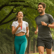 Two people jogging together in a park, both smiling. The woman wears a white top and teal leggings, while the man wears a dark athletic shirt and shorts. Trees and a path with green foliage are visible in the background, reminiscent of the scenic Ann and Roy Butler Trail featured in many Runner’s Guides. Austin Marathon Half Marathon & 5K