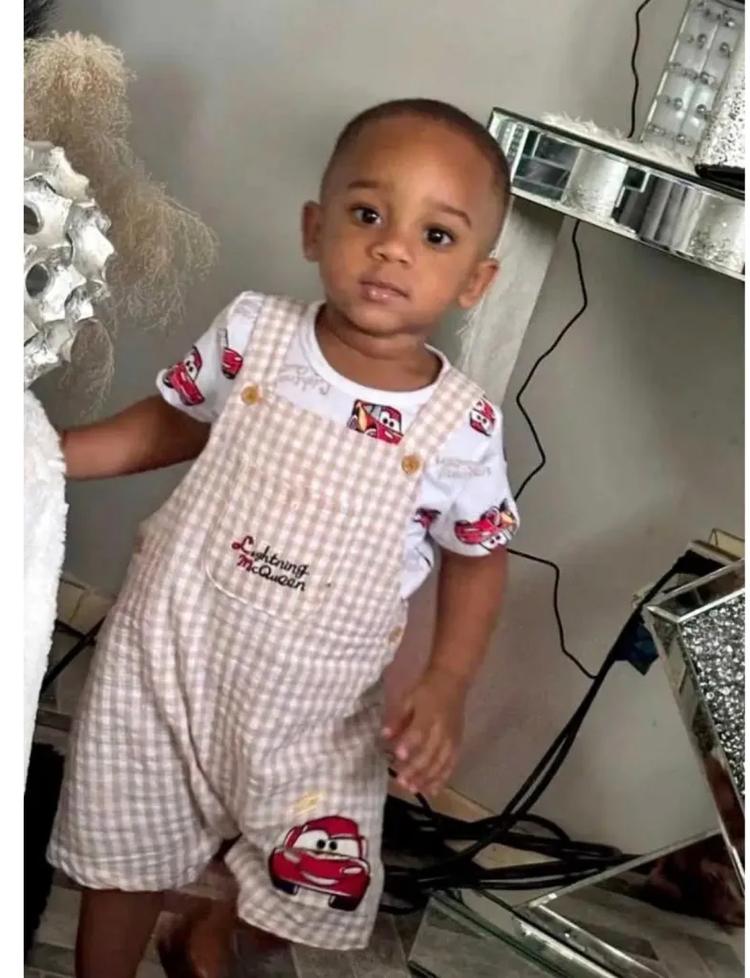 A young child wearing a white and red checkered outfit with cartoon car designs, standing in a room with various items in the background.