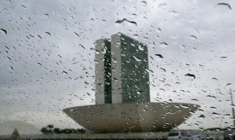 Foto colorida que mostra lente ou vidro com gotas de chuva e no fundo, o prédio do Congresso Nacional. Mais ao fundo, nuvens carregadas