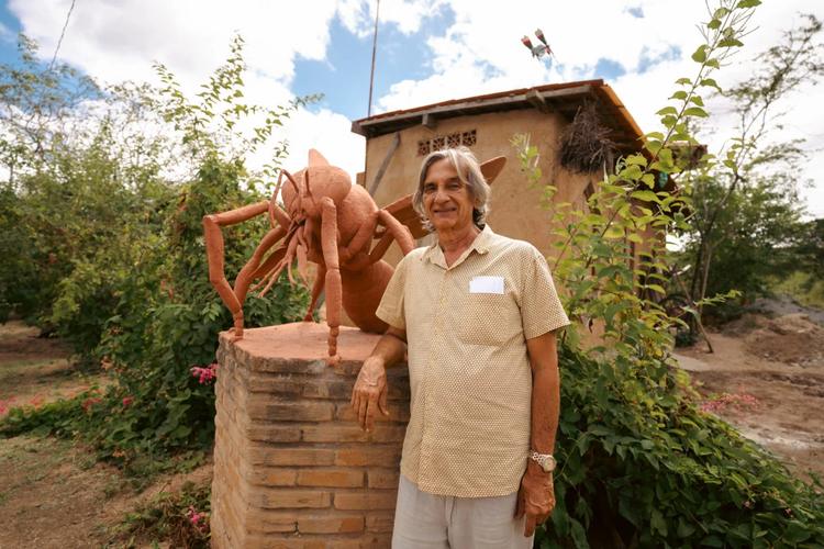 Foto colorida de Lula do Mel. Ele é um homem branco, com cabelo grisalho na altura do ombro. Ele está ao lado de uma escultura de uma abelha, no Solar das Abelhas, em Caruaru.