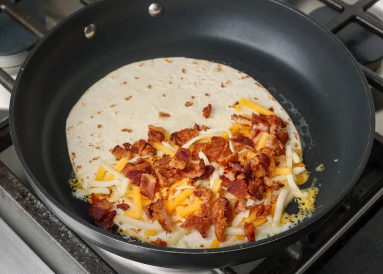 the first layer of cheese and bacon have been added to half the quesadilla in a non-stick pan