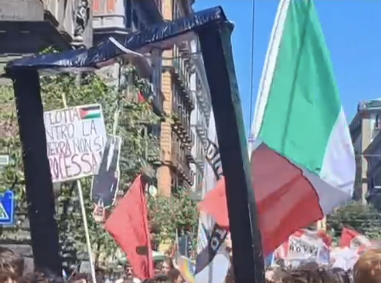 Valditara a testa in giù sotto un patibolo di cartone