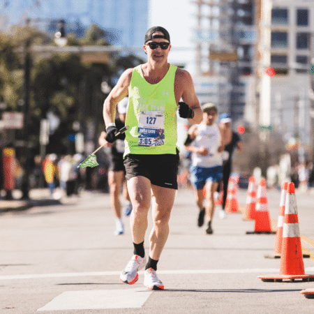 A runner in a neon green tank top, black shorts, and sunglasses competes in the Austin Marathon. He holds a small flag and wears a race bib that reads