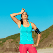 A runner in a blue tank top and pink leggings wipes sweat from her forehead while wearing earbuds and an arm-mounted phone case, taking a moment to focus on exercise recovery. Austin Marathon Half Marathon & 5K