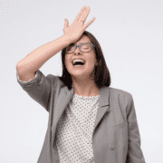 A woman wearing glasses and a gray blazer smiles with her eyes closed, touching her forehead in a classic 