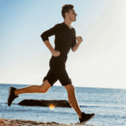 A man in black athletic wear is jogging along a sandy beach with the ocean and clear sky in the background, focusing on his marathon fueling and testing his gut training for optimal marathon nutrition. Austin Marathon Half Marathon & 5K