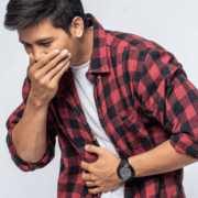 Young man in a red plaid shirt covers his mouth with one hand and holds his stomach with the other, appearing unwell or nauseous—perhaps struggling with gut training or issues related to marathon fueling—against a plain light background. Austin Marathon Half Marathon & 5K