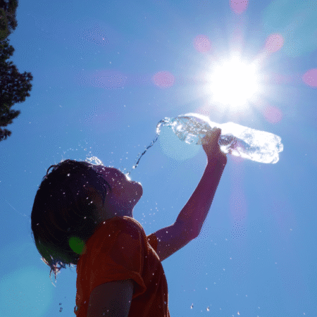 A child in an orange shirt drinks water from a plastic bottle outdoors, sunlight shining overhead and water droplets shimmering—perfect hydration strategies for hot days. Austin Marathon Half Marathon & 5K