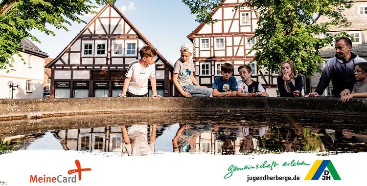  Kinder und ein erwachsener Mann am Brunnen sitzend im Hintergrund befinden sich Fachwerkhäuser. Werbung des Deutschen Jugendherbergwerks für MeineCard Plus