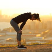 A woman in athletic clothing stands outdoors at sunset, bent over with hands on knees, appearing tired after training. A cityscape and body of water are visible in the blurred background, hinting at the importance of burnout prevention. Austin Marathon Half Marathon & 5K