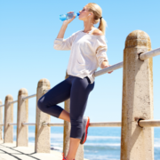 Woman in athletic wear drinks water while leaning against a seaside railing on a sunny day, earphones in, with the ocean and clear blue sky in the background—taking a mindful break to reflect on February planning and gather productivity tips for better time management. Austin Marathon Half Marathon & 5K