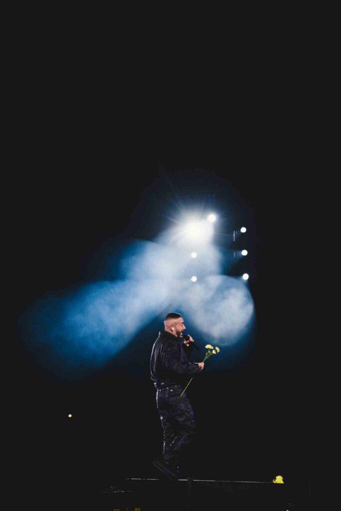 Male singer on stage under a blue spotlight, holding a microphone and a yellow flower, performing in a dark venue with fog effects.