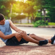 A man in athletic clothes sits on an outdoor track, holding his knee and appearing to be in pain—perhaps searching for running tips on how to handle a missed run. Trees and sunlight are visible in the background. Austin Marathon Half Marathon & 5K