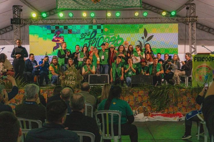 Foto colorida do palco da 32° Feicoop, durante cerimônia de abertura do evento. Na imagem, aparecem diversas pessoas da organização reunidas no palco. Evento discute impacto da crise climática na economia solidária