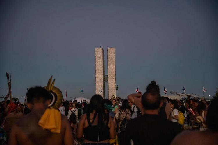 Foto colorida de indígenas de costas durante o Acampamento Terra Livre de 2023, em Brasília. Ao fundo, prédio do Congresso