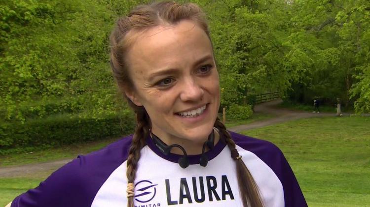 Laura Aiken, a woman with long, fair hair tied back in two plaits, speaks to a BBC TV crew at the Stormont estate.  She is wearing a two-tone sports top with a white chest and purple sleeves, and a set of black headphones. 