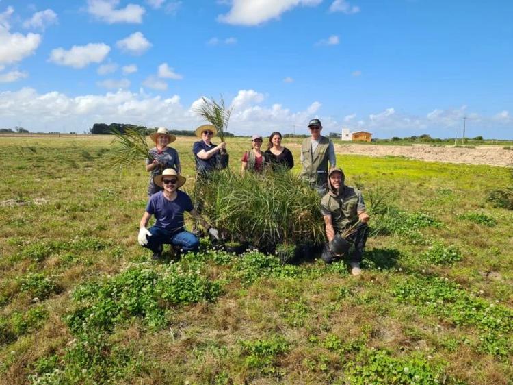Foto colorida da família de Bruna e equipe do Herbário BLA durante plantio de mudas de butiá em Santa Vitória do Palmar