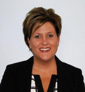 A woman wearing a white-and-black patterned shirt and a black blazer smiles at the camera. A plain white background is behind her.