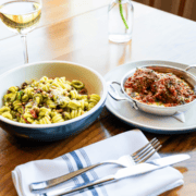 A bowl of pesto pasta with pine nuts alongside a small pan of meatballs in red sauce, with a glass of white wine and cutlery on a wooden table, perfect for pre-run pasta. Austin Marathon Half Marathon & 5K