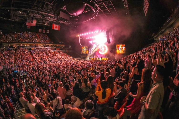 Crowd gathers in a packed arena for a concert, bright stage lights and large screens behind the performers.