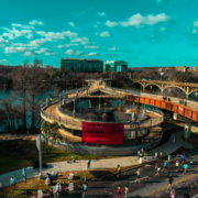 People race on a road near a spiral pedestrian bridge with a large pink sign saying “ASK YOUR HEROES FOR FORGIVENESS, NOT PERMISSION.” Modern buildings and a river are in the background under a blue sky—the perfect setting for morning runs in Austin or discovering some of the best running routes Austin has to offer. Austin Marathon Half Marathon & 5K