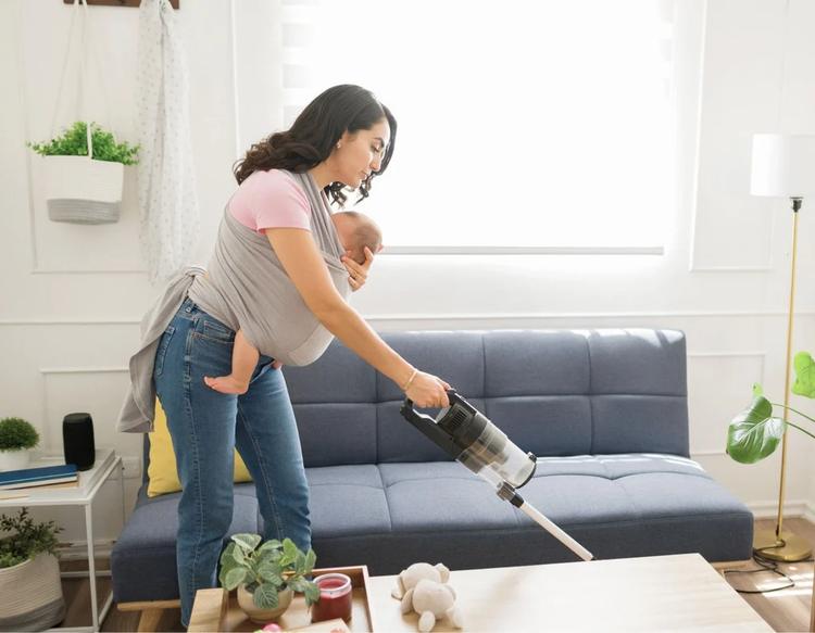A mother babywearing her infant in a supportive wrap while vacuuming and organizing a sunlit living room for spring cleaning.