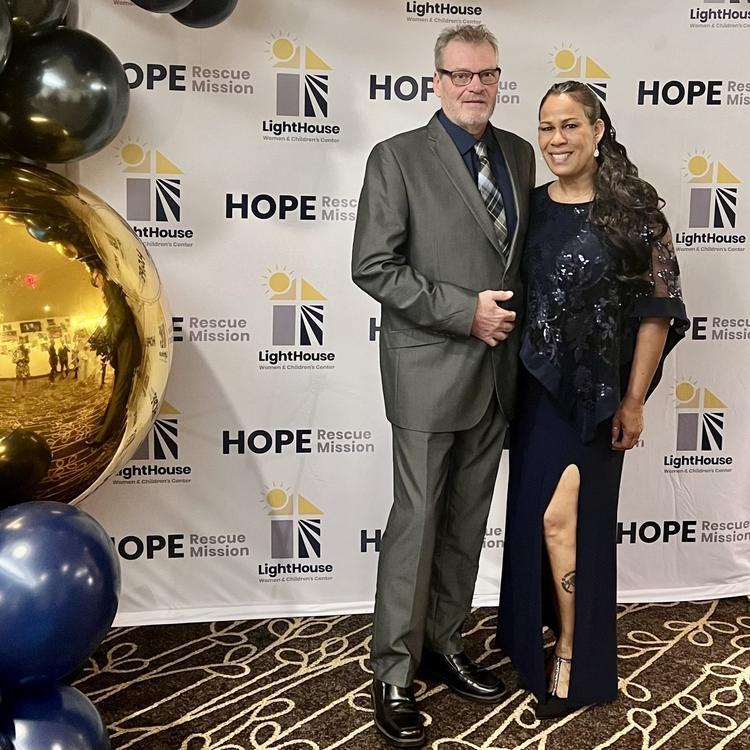 A man and a woman pose for a picture in front of a large banner that has logos for two organizations: Hope Rescue Mission and Lighthouse. There are gold, black and grey balloons hanging around the banner. The man and the woman are wearing formal clothes.