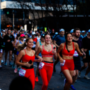Three women in matching red athletic outfits smile and run together during Race Weekend, surrounded by other runners on a city street. The atmosphere is energetic and lively—perfect for an unforgettable Austin vacation. Austin Marathon Half Marathon & 5K