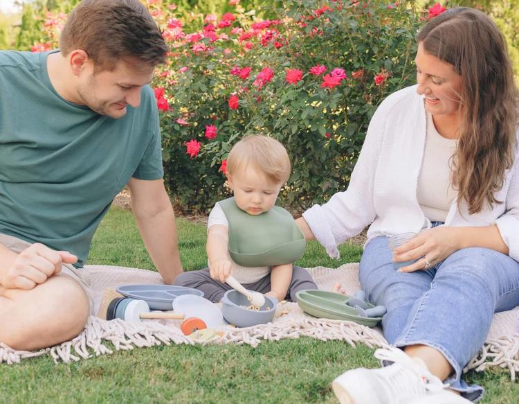Family watching baby self-feed during a sunny summer picnic outdoors.