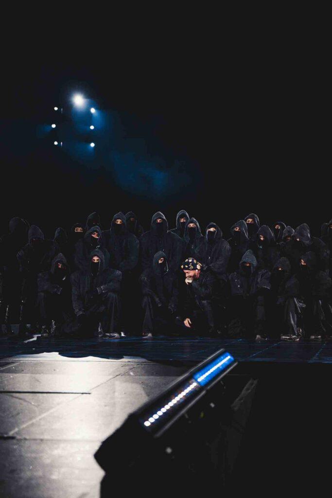 A line of people in dark hooded robes on a dim stage, blue spotlights overhead and stage equipment visible at the front.