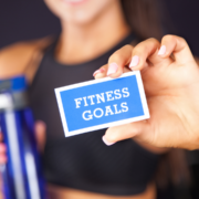 A person in athletic wear holds a blue card reading “FITNESS GOALS” and a blue water bottle, emphasizing a focus on fitness, healthy living, and the importance of a Mid-Year Fitness Check. Austin Marathon Half Marathon & 5K