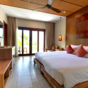 A cozy hotel room perfect for your Austin Vacation, featuring a large bed with four red pillows, wooden decor, a ceiling fan, a sitting area, and glass doors leading to a sunny patio surrounded by greenery. Austin Marathon Half Marathon & 5K