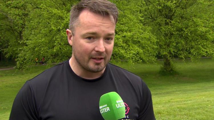 Graham Rennie, a man with short, fair hair, talks to a BBC TV crew in the grounds of Stormont.  He is wearing a black crew-neck t-shirt and is talking into a green microphone.  There is a lawned area covered in mature trees in the background.