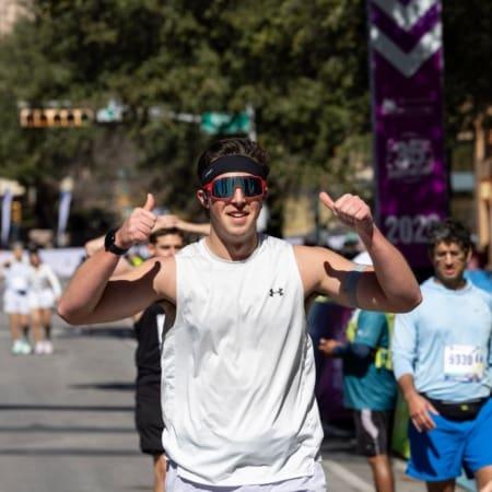 A man in a white athletic shirt and sunglasses smiles and gives two thumbs up while running in a race, embracing what's next. Other runners and race banners are visible in the background on a sunny day. Austin Marathon Half Marathon & 5K
