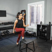 Woman in athletic clothing doing a step-up exercise on a black platform in a home gym, surrounded by dumbbells and fitness equipment—perfect for essential workouts that support marathon longevity. Austin Marathon Half Marathon & 5K