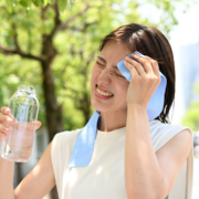 A woman outdoors wipes sweat from her forehead with a blue cloth while holding a bottle of water. She squints, appearing hot and uncomfortable beneath the sun—determined to stay on track with the Summer Consistency Challenge. Austin Marathon Half Marathon & 5K