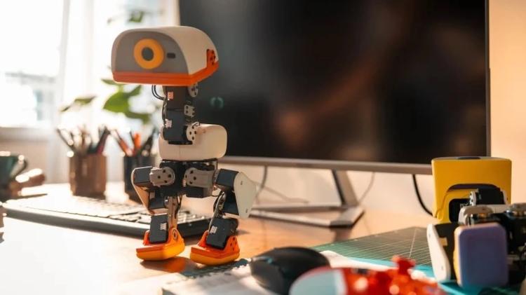 A small orange-and-white humanoid robot stands on a desk in front of a computer monitor.