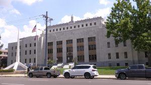 The Hinds County Courthouse stands behind a monument and a row of parked cars. The words “Hinds County Court House” are carved into the front of the building.