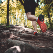 A person wearing black shorts and bright red running shoes jogs uphill on a rugged, root-covered forest trail, surrounded by tall trees and dappled sunlight, striving to build running distance while enjoying nature and avoiding burnout. Austin Marathon Half Marathon & 5K