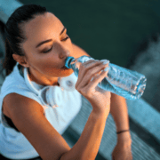A woman in sportswear with headphones around her neck drinks water from a plastic bottle outdoors, possibly after exercising and checking off items from her Running Checklist. Austin Marathon Half Marathon & 5K