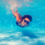 A woman wearing sunglasses and a swimsuit swims underwater in a pool, smiling toward the camera as sunlight reflects off the water—enjoying a moment to refuel and recharge, perfect for post-run Austin recovery. Austin Marathon Half Marathon & 5K