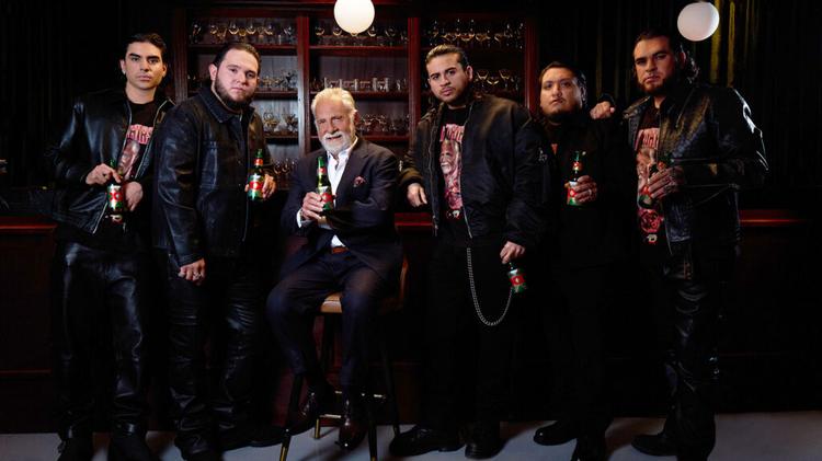 Older man in a suit seated on a bar stool, surrounded by six men in black leather jackets, all holding beer bottles in a dim bar setting.