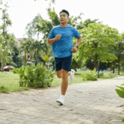 Man in blue shirt and shorts jogging on a paved path in a park, surrounded by green trees and plants on a sunny day—perfect for practicing gut training as part of his marathon plan. Austin Marathon Half Marathon & 5K