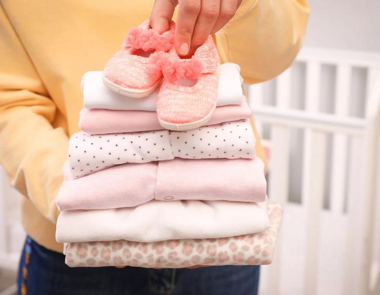 Woman holding stack of girl's clothes and booties indoors, closeup.