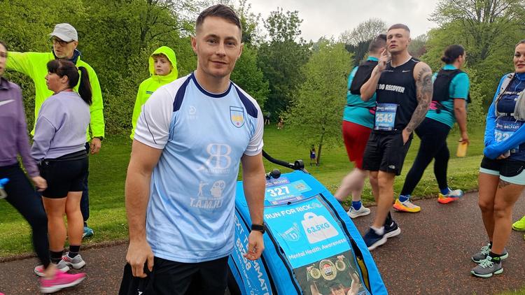 Ryan Burnett, a man with short, fair hair, poses at the start line with a weighted trolley. He is wearing a pale blue and navy t-shirt with white sleeves.  The trolley  has a blue cover with a photo of the late Ricky Hatton holding up a boxing belt above his head in celebration.  The text on the trolley reads: 