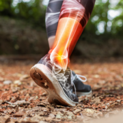 A person in athletic wear walking on a leaf-covered trail, with a digital illustration overlay highlighting pain in the ankle area, perhaps due to intense marathon training or following demanding training plans. Austin Marathon Half Marathon & 5K