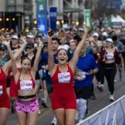 Three smiling women in red athletic outfits with race bibs cheer and raise their arms while running together as a team in a large outdoor charity race; many other runners are behind them on the city street. Austin Marathon Half Marathon & 5K