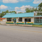 A small restaurant named “Juan in a Million” with blue trim and yellow benches sits on a quiet street under a partly cloudy sky, surrounded by trees and a metal fence—a popular spot for East Austin running clubs to gather and refuel with post-run tacos. Austin Marathon Half Marathon & 5K